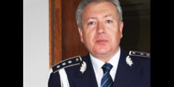 Middle-aged man in formal max blue military uniform with epaulets, medals, and striped tie, standing indoors.