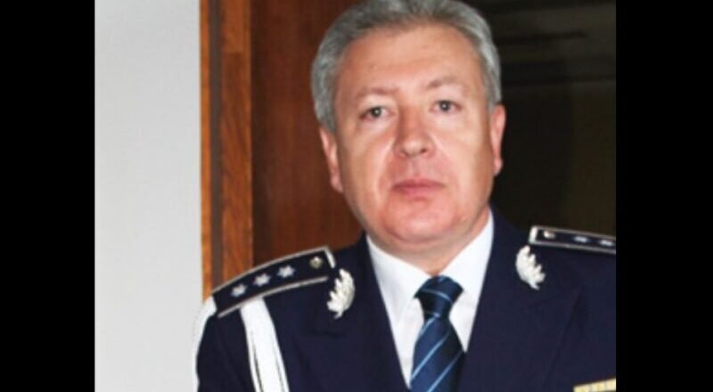 Middle-aged man in formal max blue military uniform with epaulets, medals, and striped tie, standing indoors.