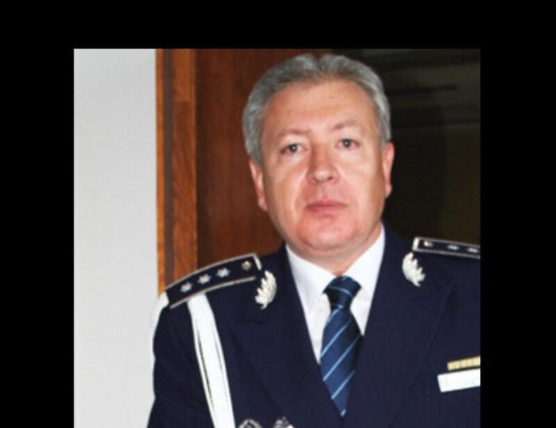 Middle-aged man in formal max blue military uniform with epaulets, medals, and striped tie, standing indoors.