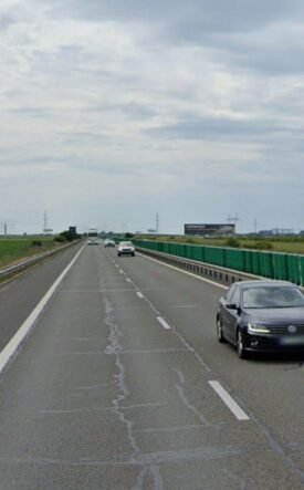 Black sedan in the right lane of a divided highway with green guardrails and open fields on both sides under a cloudy sky.