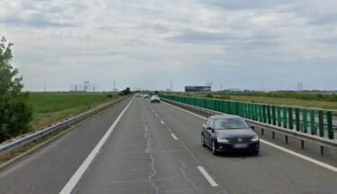 Black sedan in the right lane of a divided highway with green guardrails and open fields on both sides under a cloudy sky.