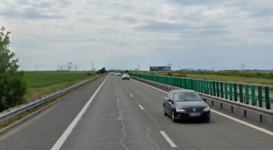 Black sedan in the right lane of a divided highway with green guardrails and open fields on both sides under a cloudy sky.