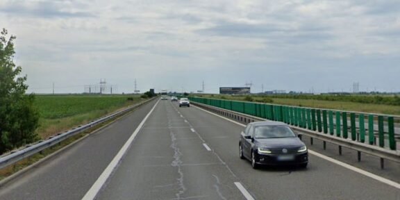 Black sedan in the right lane of a divided highway with green guardrails and open fields on both sides under a cloudy sky.
