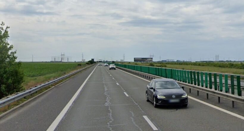 Black sedan in the right lane of a divided highway with green guardrails and open fields on both sides under a cloudy sky.