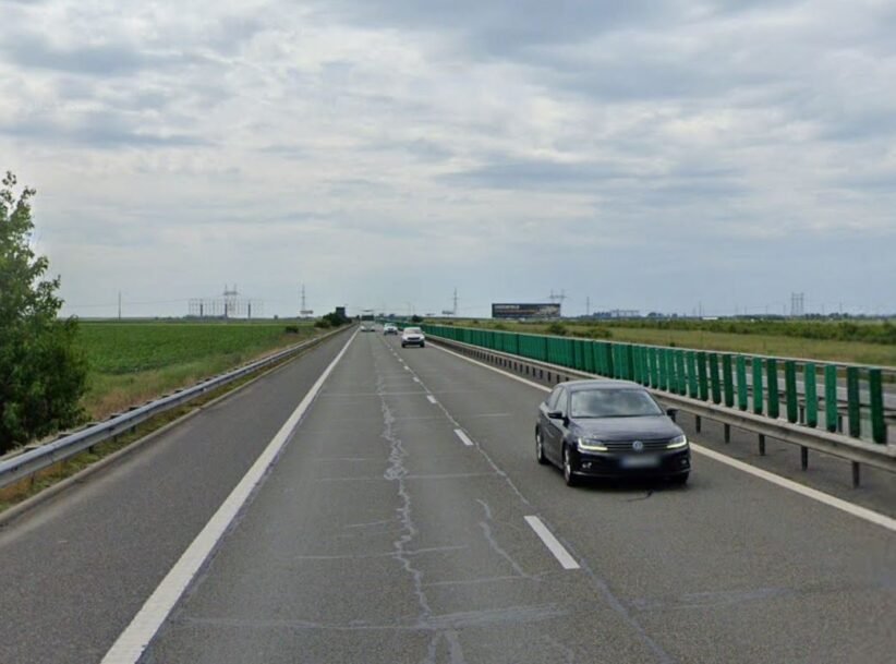 Black sedan in the right lane of a divided highway with green guardrails and open fields on both sides under a cloudy sky.