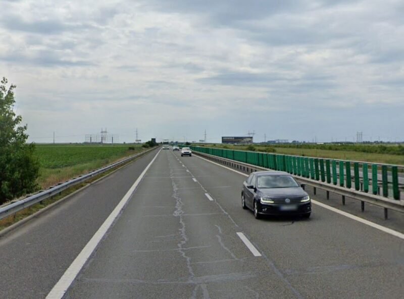 Black sedan in the right lane of a divided highway with green guardrails and open fields on both sides under a cloudy sky.