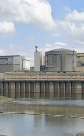 Industrial facility with large storage tanks and a dam along a calm river under a partly cloudy sky.