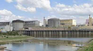 Industrial facility with large storage tanks and a dam along a calm river under a partly cloudy sky.