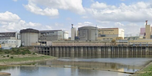 Industrial facility with large storage tanks and a dam along a calm river under a partly cloudy sky.