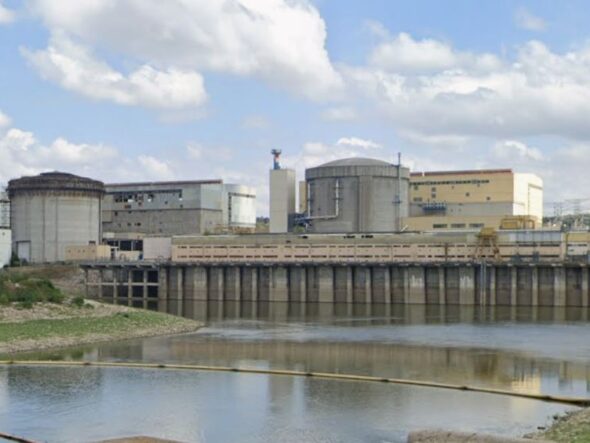 Industrial facility with large storage tanks and a dam along a calm river under a partly cloudy sky.