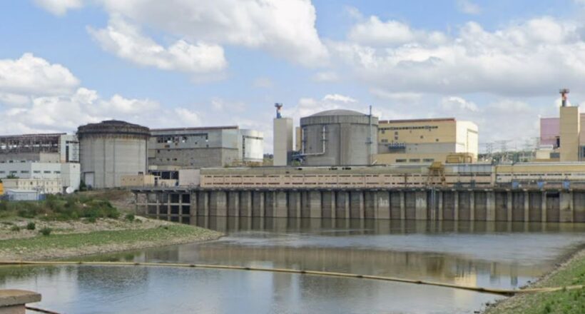 Industrial facility with large storage tanks and a dam along a calm river under a partly cloudy sky.