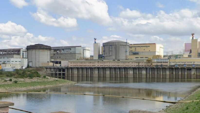 Industrial facility with large storage tanks and a dam along a calm river under a partly cloudy sky.