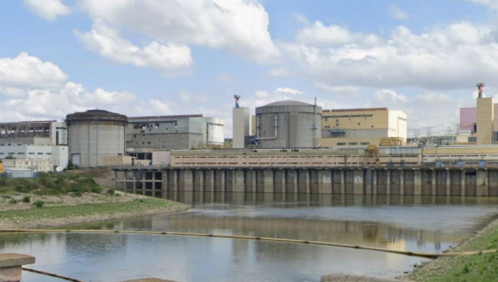 Industrial facility with large storage tanks and a dam along a calm river under a partly cloudy sky.