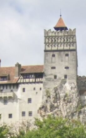 Medieval fortress atop a rocky outcrop with crenellated towers and red-tiled roofs, surrounded by trees.