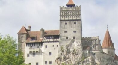 Medieval fortress atop a rocky outcrop with crenellated towers and red-tiled roofs, surrounded by trees.