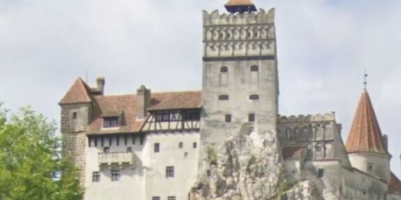 Medieval fortress atop a rocky outcrop with crenellated towers and red-tiled roofs, surrounded by trees.
