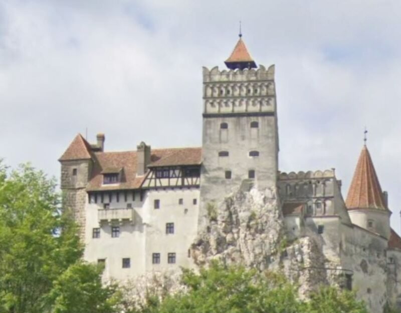 Medieval fortress atop a rocky outcrop with crenellated towers and red-tiled roofs, surrounded by trees.
