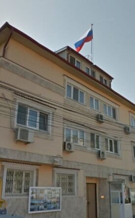 Beige three-story building with window air conditioners, a Russian flag flying from the roof, and a narrow street with a streetlight nearby.