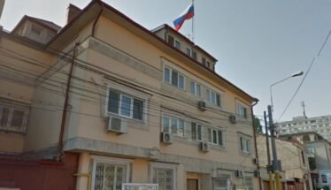 Beige three-story building with window air conditioners, a Russian flag flying from the roof, and a narrow street with a streetlight nearby.