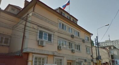 Beige three-story building with window air conditioners, a Russian flag flying from the roof, and a narrow street with a streetlight nearby.