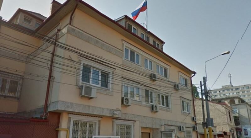 Beige three-story building with window air conditioners, a Russian flag flying from the roof, and a narrow street with a streetlight nearby.