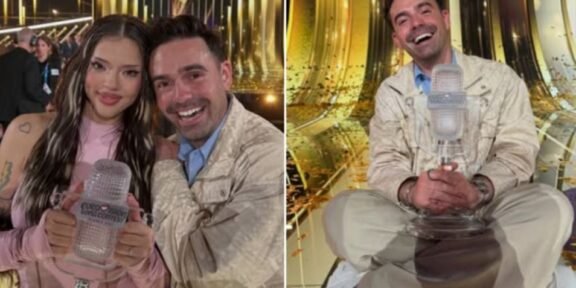 Woman in pink holds a clear Eurovision trophy while a smiling man stands beside her in a glittering backstage area.