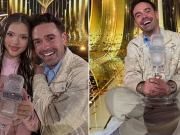 Woman in pink holds a clear Eurovision trophy while a smiling man stands beside her in a glittering backstage area.