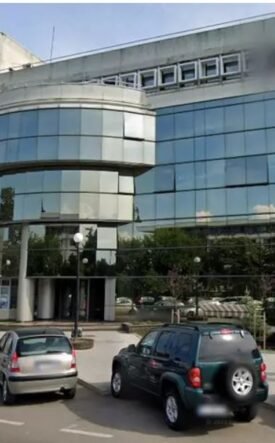 Front view of a modern office building with a curved glass façade and a row of parked cars in a small lot.