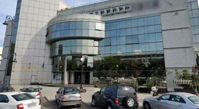 Front view of a modern office building with a curved glass façade and a row of parked cars in a small lot.