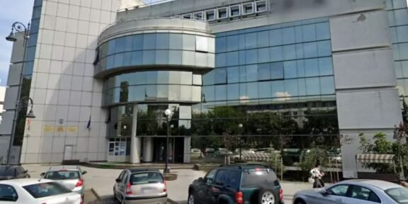 Front view of a modern office building with a curved glass façade and a row of parked cars in a small lot.