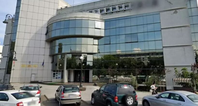 Front view of a modern office building with a curved glass façade and a row of parked cars in a small lot.