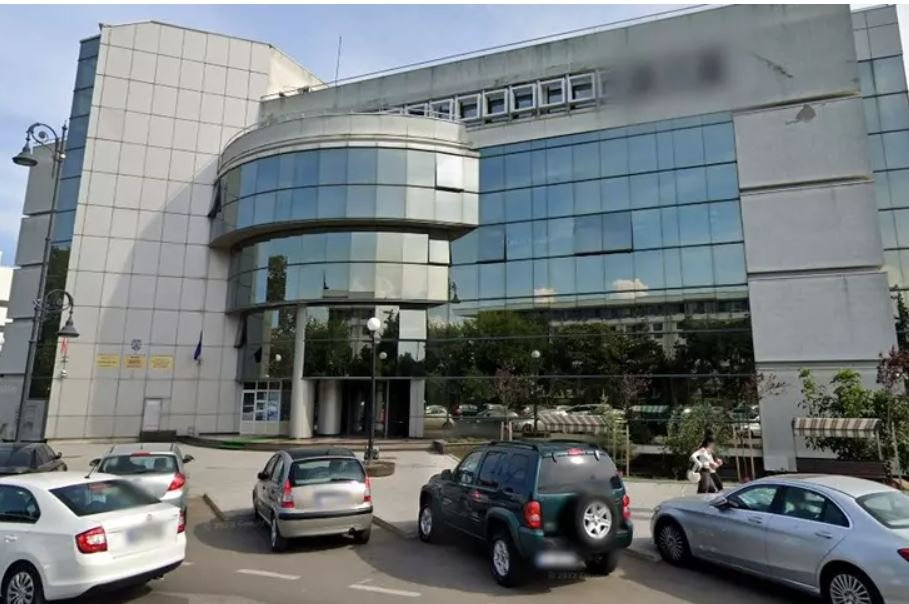 Front view of a modern office building with a curved glass façade and a row of parked cars in a small lot.