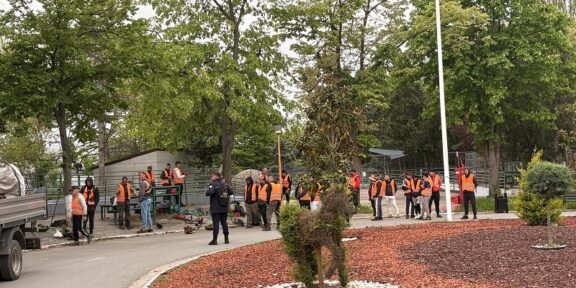 Group of workers in orange safety vests gathered around equipment in a park-like outdoor worksite with trees nearby.