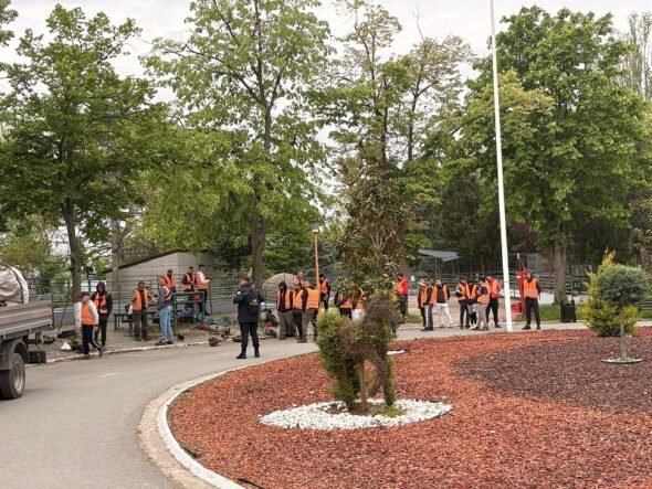 Group of workers in orange safety vests gathered around equipment in a park-like outdoor worksite with trees nearby.