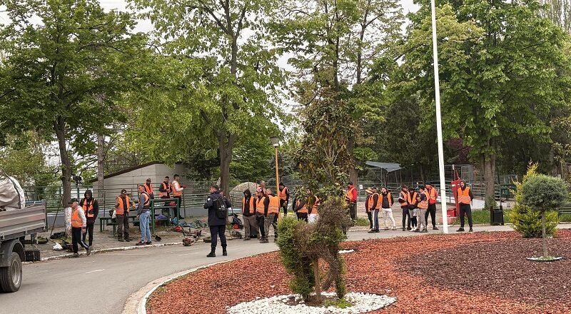 Group of workers in orange safety vests gathered around equipment in a park-like outdoor worksite with trees nearby.