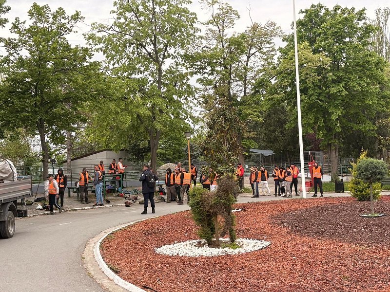 Group of workers in orange safety vests gathered around equipment in a park-like outdoor worksite with trees nearby.
