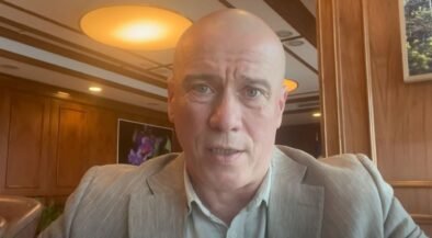 Close-up of a bald man in a gray suit speaking to the camera in a wood-paneled room.