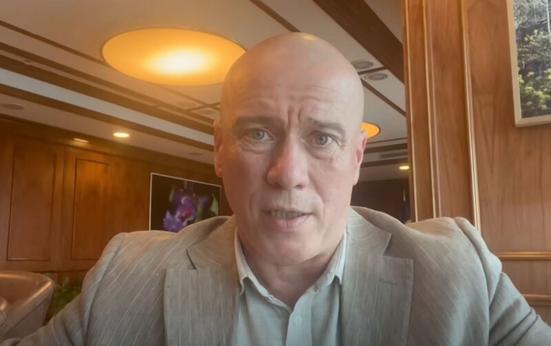 Close-up of a bald man in a gray suit speaking to the camera in a wood-paneled room.