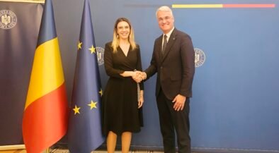 Two formally dressed individuals shake hands in front of a blue backdrop that reads 'GUVERNUL ROMÂNIEI', flanked by Romanian and European Union flags.
