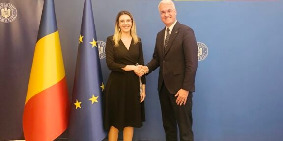 Two formally dressed individuals shake hands in front of a blue backdrop that reads 'GUVERNUL ROMÂNIEI', flanked by Romanian and European Union flags.
