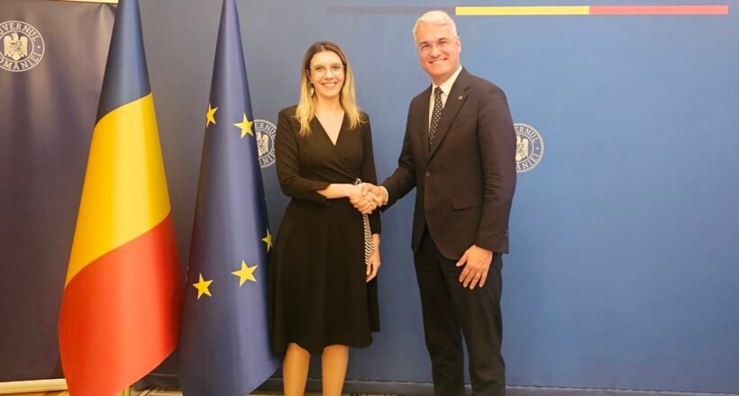 Two formally dressed individuals shake hands in front of a blue backdrop that reads 'GUVERNUL ROMÂNIEI', flanked by Romanian and European Union flags.