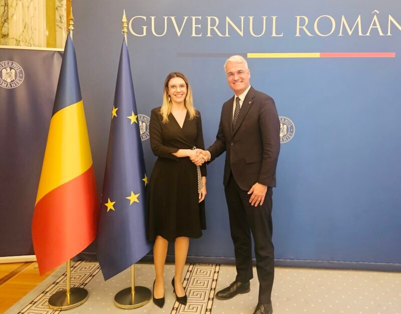 Two formally dressed individuals shake hands in front of a blue backdrop that reads 'GUVERNUL ROMÂNIEI', flanked by Romanian and European Union flags.