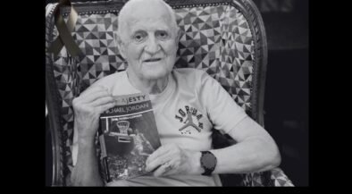 Older man with a white T-shirt sits in a patterned chair, holding a Michael Jordan basketball book and wearing a watch; a black ribbon is pinned to the chair in the background.