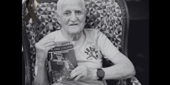 Older man with a white T-shirt sits in a patterned chair, holding a Michael Jordan basketball book and wearing a watch; a black ribbon is pinned to the chair in the background.
