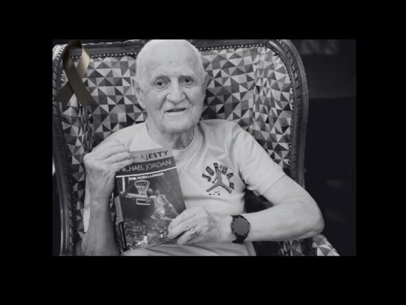 Older man with a white T-shirt sits in a patterned chair, holding a Michael Jordan basketball book and wearing a watch; a black ribbon is pinned to the chair in the background.