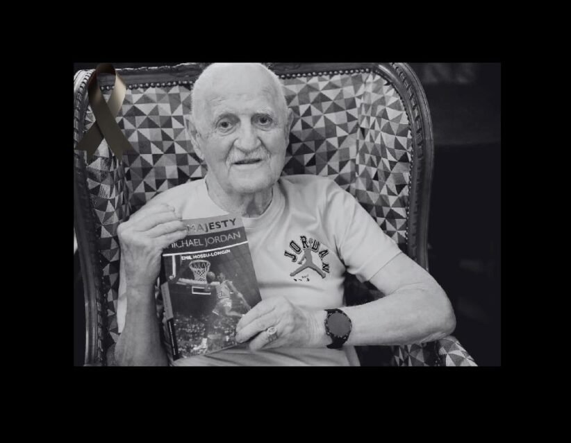 Older man with a white T-shirt sits in a patterned chair, holding a Michael Jordan basketball book and wearing a watch; a black ribbon is pinned to the chair in the background.