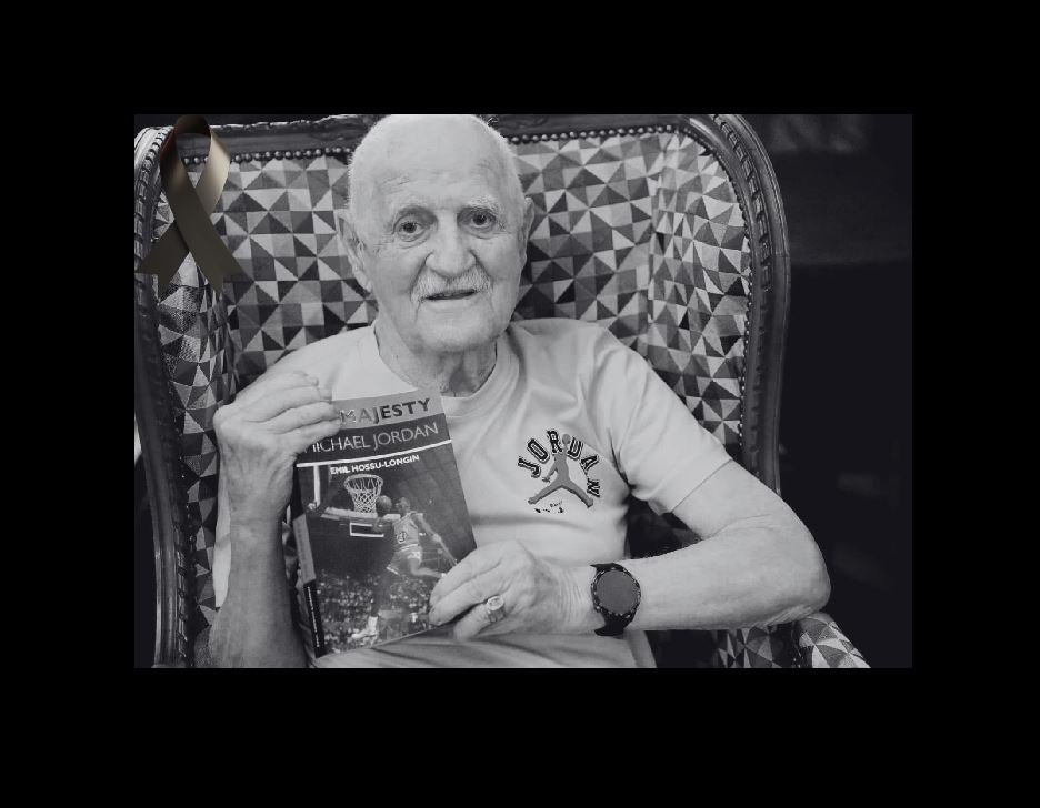 Older man with a white T-shirt sits in a patterned chair, holding a Michael Jordan basketball book and wearing a watch; a black ribbon is pinned to the chair in the background.