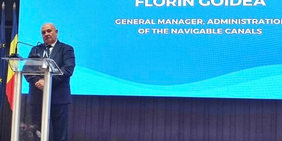 Man in a dark suit giving a speech at a clear podium on a stage, with a large blue presentation screen behind him and a Romanian flag visible on the left