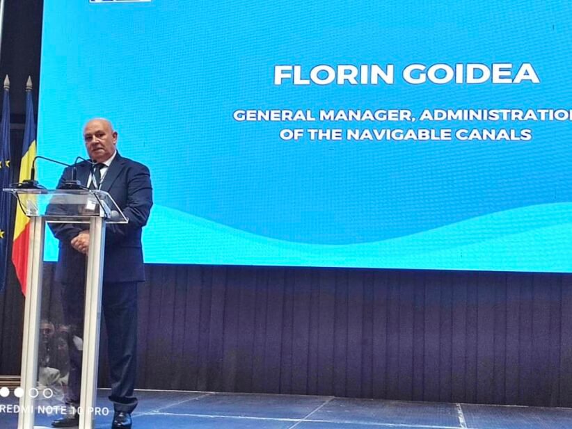 Man in a dark suit giving a speech at a clear podium on a stage, with a large blue presentation screen behind him and a Romanian flag visible on the left