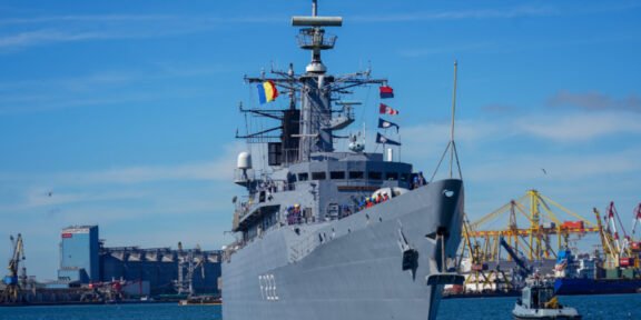 Gray naval destroyer docked at a harbor with cranes and a bright blue sky behind it, flags on the mast visible at the bow.
